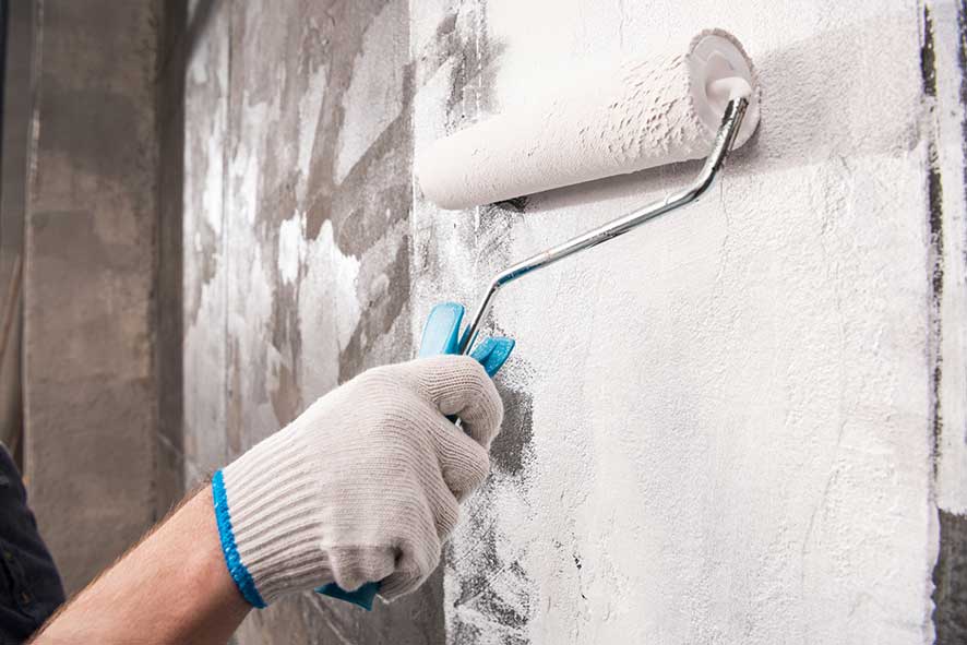 Hand with work glove painting a concrete wall with white paint and a roller brush.